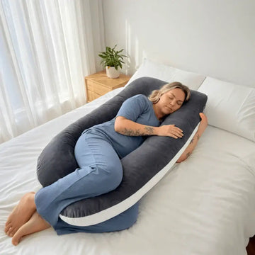Woman sleeping comfortably on her side wrapped around The Hug pregnancy pillow on a white bed in a bright minimalist bedroom