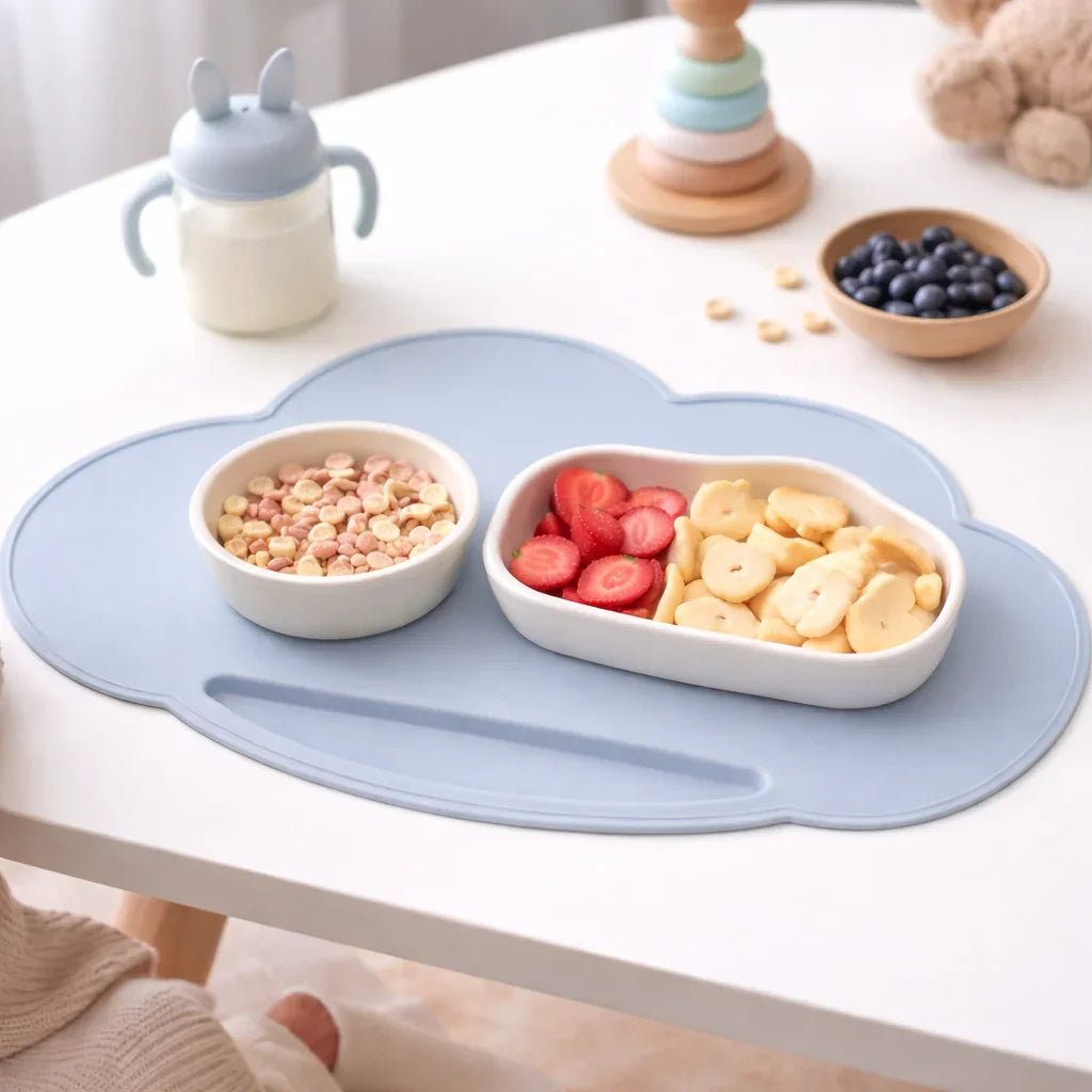 Blue cloud-shaped silicone placemat with cereal, sliced strawberries, bananas, toddler cup on table